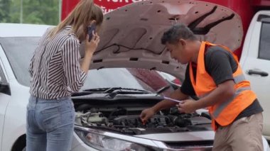 Unhappy Asian female car owner standing in the rain at her car and talking on her phone while the car insurance agent inspecting her car for the insurance claim report