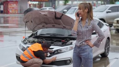 Unhappy Asian female car owner standing in the rain at her car and talking on her phone while the car insurance agent inspecting her car for the insurance claim report