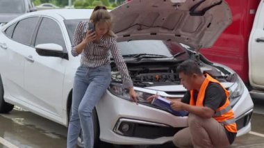 Unhappy Asian female car owner standing in the rain at her car and talking on her phone while the car insurance agent inspecting her car for the insurance claim report
