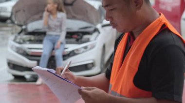 Close up of a male car insurance agent filling in a car repair claim form with female car owner talking on her phone near her car