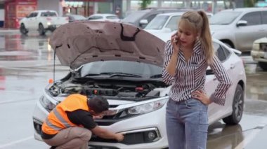 Unhappy Asian female car owner standing in the rain at her car and talking on her phone while the car insurance agent inspecting her car for the insurance claim report