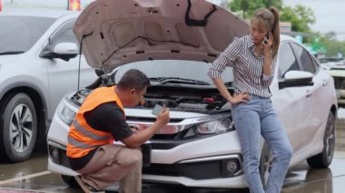 Unhappy Asian female car owner standing in the rain at her car and talking on her phone while the car insurance agent inspecting her car for the insurance claim report