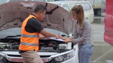 Unhappy Asian woman signing the insurance claim report form from a male car insurance agent in front of her car on a raining day