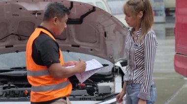 Unhappy Asian woman signing the insurance claim report form from a male car insurance agent in front of her car on a raining day