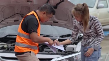 Unhappy Asian woman signing the insurance claim report form from a male car insurance agent in front of her car on a raining day