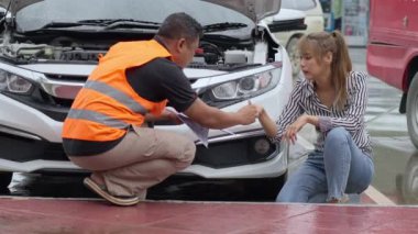 Unhappy Asian woman signing the insurance claim report form from a male car insurance agent in front of her car on a raining day