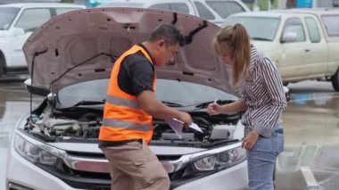 Unhappy Asian woman signing the insurance claim report form from a male car insurance agent in front of her car on a raining day