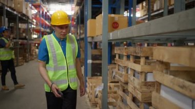 Male warehouse staff wearing safety vest and yellow hardhat using a handheld scanning device to check the inventory inside the warehouse