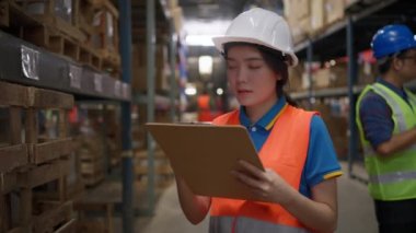 Young female warehouse staff wearing safety vest and hard hat holding a clipboard while checking the product stock inventory in the store warehouse