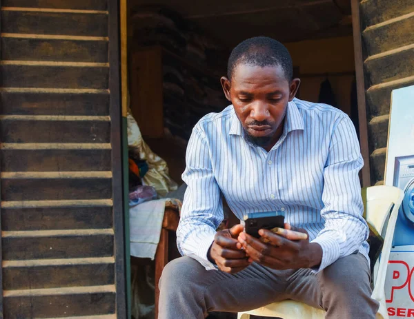 Afrikalı bir Nijeryalı işadamı, tüccar ya da girişimci dükkanının önünde oturuyor ve elinde akıllı bir telefon kullanıyor.