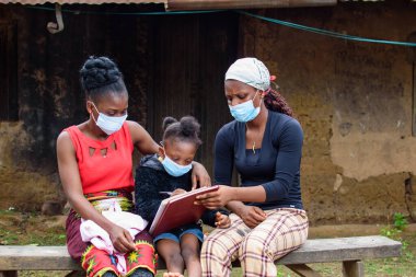 İki Afrikalı kadın, burun maskeli anne ve öğretmen, köyün çamurlu evinin dışında küçük bir kızla birlikte oturuyorlar, okulunda, kariyerinde ve eğitiminde mükemmellik için çalışmalarına yardım ediyorlar.