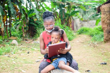 Bir Afrikalı anne ya da öğretmen köyün çamurlu evinin önünde, burun maskeli bir kız çocuğuna okulunda, kariyerinde ve eğitiminde mükemmellik için yardım ediyor.