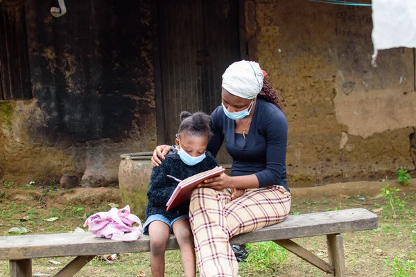 An African mother or teacher outside a village mud house with nose mask, helping a girl child with her studies for excellence in her school, career and education