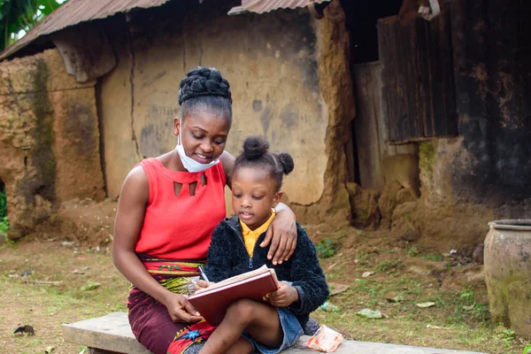 An African mother or teacher outside a village mud house with nose mask, helping a girl child with her studies for excellence in her school, career and education