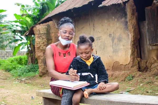 An African mother or teacher outside a village mud house with nose mask, helping a girl child with her studies for excellence in her school, career and education