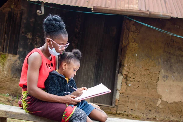 An African mother or teacher outside a village mud house with nose mask, helping a girl child with her studies for excellence in her school, career and education