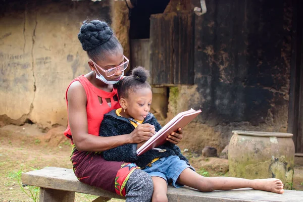 An African mother or teacher outside a village mud house with nose mask, helping a girl child with her studies for excellence in her school, career and education