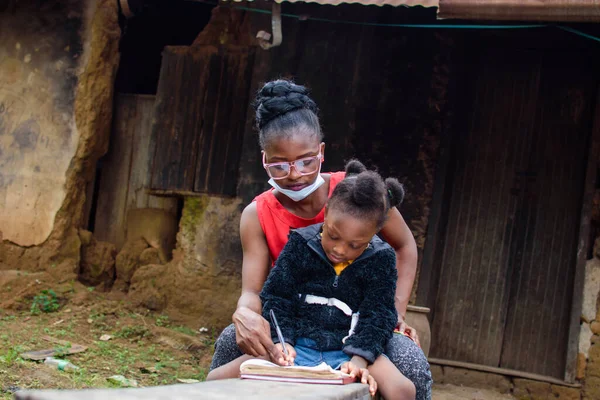 An African mother or teacher outside a village mud house with nose mask, helping a girl child with her studies for excellence in her school, career and education