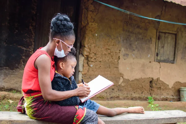 An African mother or teacher outside a village mud house with nose mask, helping a girl child with her studies for excellence in her school, career and education