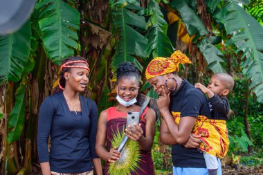 Three female africans standing and looking at a smart phone with different expression on a banana farm while one of the women have a baby on her back
