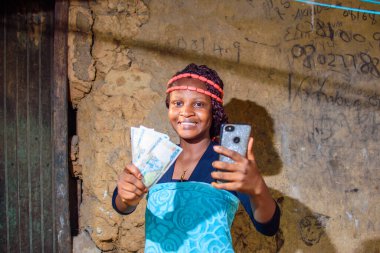 A female african standing outside a village mud house, holding a smart phone and multiple Nigerian cash, money, Naira notes or currency in her hand