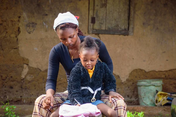 An African mother, guardian or teacher outside a village mud house, helping a girl child or student with her studies for excellence in her school, career and education