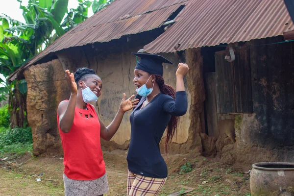 An African woman with nose mask joyfully celebrates with a female graduand or student of her success in education in front of a village mud house