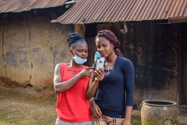 Two happy african women or ladies wearing nose mask, stands outside a village mud house, holding and looking into a smart phone with them