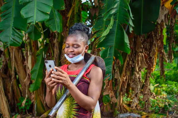 A female african farmer with nose mask and a farming hoe on her shoulder happily looks into a smart phone she\'s holding in a banana farm or plantation