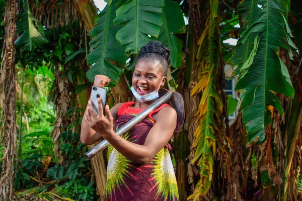A female african farmer with nose mask and a farming hoe on her shoulder happily looks into a smart phone she's holding in a banana farm or plantation