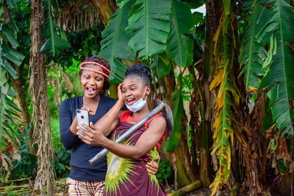 A female african farmer with nose mask and a farming hoe on her shoulder happily looks into a smart phone with a female friend in a banana farm or plantation