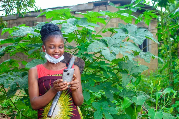 A female african farmer with nose mask and a farming hoe on her shoulder happily looks into a smart phone she is holding in a house farm or garden