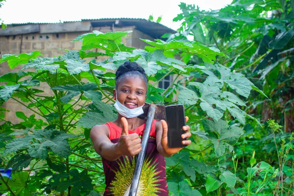 A female african farmer with nose mask and a farming hoe on her shoulder happily shows and points to the smart phone she is holding in a farm or garden