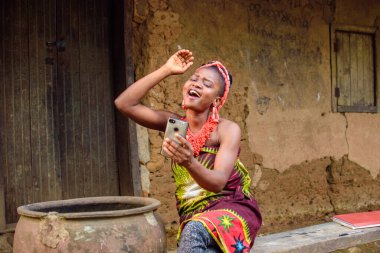 Kafasında boncuklar olan mutlu bir Afrikalı kadın ya da kadın, büyük su kabağının yanında oturuyor, heyecanla bir köy çamur evinin önünde akıllı bir telefon tutuyor.