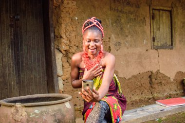 Kafasında boncuklar olan mutlu bir Afrikalı kadın ya da kadın, büyük su kabağının yanında oturuyor, heyecanla bir köy çamur evinin önünde akıllı bir telefon tutuyor.
