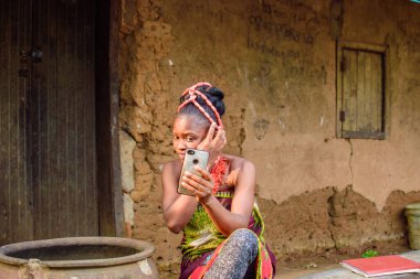 A happy African lady or woman with beads on her head, sitting beside big water calabash, excitedly holding a smart phone outside a village mud house