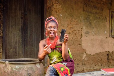 A happy African lady or woman with beads on her head, sitting beside big water calabash, excitedly pointing to a smart phone outside a village mud house clipart