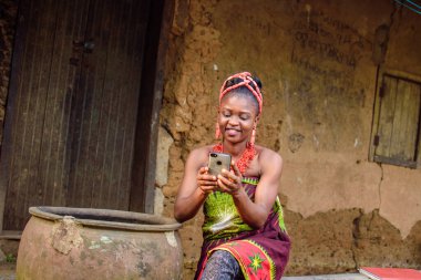 Kafasında boncuklar olan mutlu bir Afrikalı kadın ya da kadın, büyük su kabağının yanında oturuyor, heyecanla bir köy çamur evinin önünde akıllı bir telefon tutuyor.