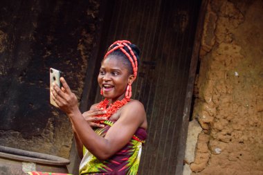 Kafasında boncuklar olan mutlu bir Afrikalı kadın ya da kadın, büyük su kabağının yanında oturuyor, heyecanla bir köy çamur evinin önünde akıllı bir telefon tutuyor.
