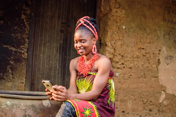 A happy African lady or woman with beads on her head, sitting beside big water calabash, excitedly holding a smart phone outside a village mud house