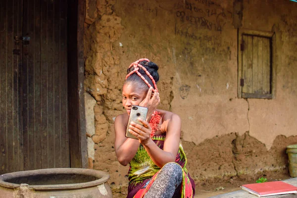 Kafasında boncuklar olan mutlu bir Afrikalı kadın ya da kadın, büyük su kabağının yanında oturuyor, heyecanla bir köy çamur evinin önünde akıllı bir telefon tutuyor.