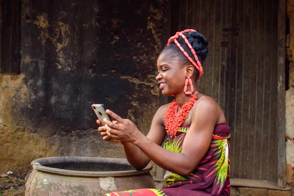 A happy African lady or woman with beads on her head, sitting beside big water calabash, excitedly holding a smart phone outside a village mud house