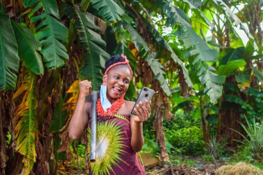 Burun maskesi takan Afrikalı bir kadın çiftçi, yerel boncuklar ve omzundaki bir çiftlik çapası muz çiftliği ya da çiftliğindeki akıllı bir telefona mutlu bir şekilde bakar.