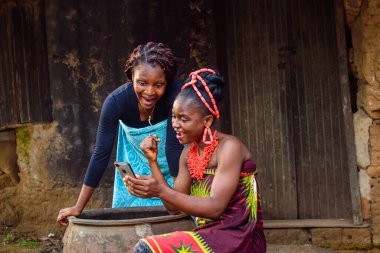 Two female Africans happily looking into the smart phone outside a village mud house that have a big water calabash clipart