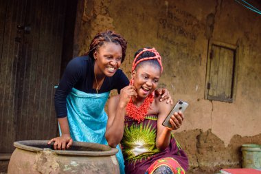 İki dişi Afrikalı, büyük bir su kabağı olan köy çamurlu evinin dışındaki akıllı telefona mutlulukla bakıyor.