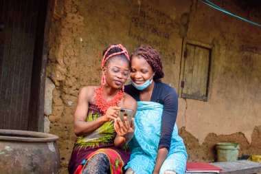 An African woman using glasses with a friend using nose mask happily look into a smart phone outside a village mud house that has a big water calabash