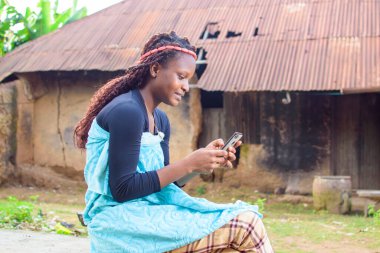 A happy woman or lady sitting and using a smart phone as she sits outside a village mud house