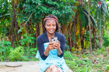 A happy woman or lady sitting and using a smart phone as she sits in a farm with banana trees behind her