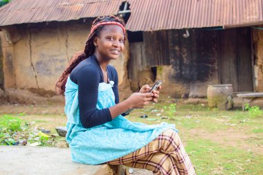 Köyün çamurlu evinin önünde oturan mutlu bir kadın ya da kadın akıllı bir telefon kullanıyor.