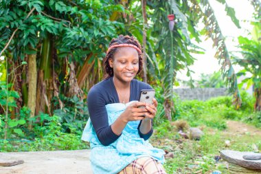 A happy woman or lady sitting and using a smart phone as she sits in a farm with banana trees behind her